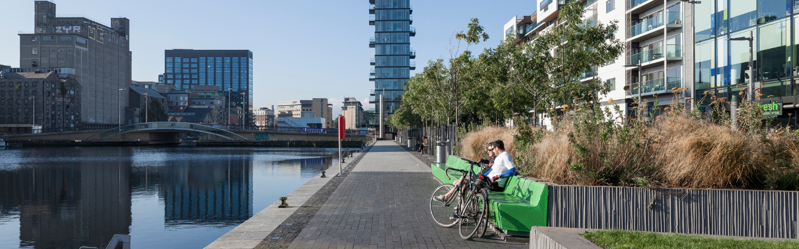 A stone-lined walkway sits next to a river, and in the distance, there are skyscrapers. There are several neon green benches along the walkway, where two pedestrians are sitting. 